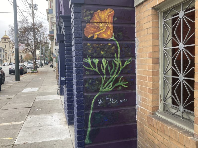 A mural of a California poppy with green stem and leaves is painted on a dark purple wall next to a sidewalk, signed "Dan 2022.