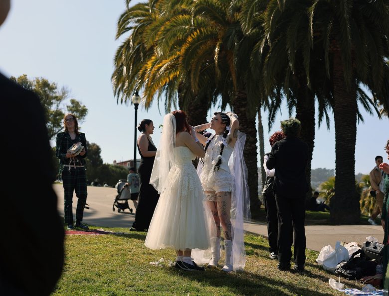 Two people in wedding attire stand facing each other outdoors by palm trees, surrounded by guests, with one adjusting the other's veil.