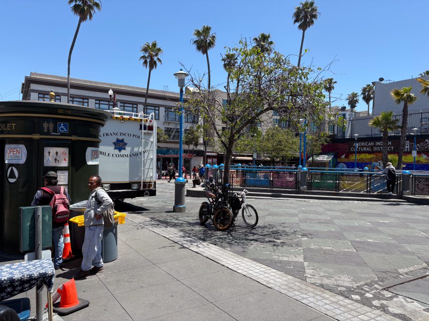 A city street scene with a public restroom, a sanitation truck, a few people, bicycles, palm trees, and colorful buildings under a clear blue sky.