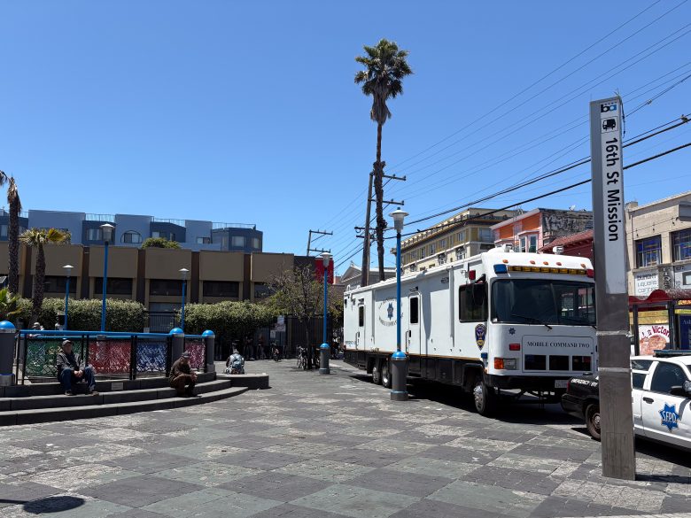 A mobile command truck is parked near the 16th St Mission BART station entrance on a sunny day, with people sitting and walking nearby.