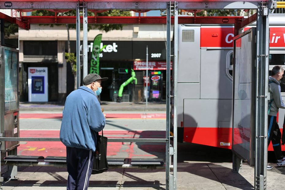 An elderly person wearing a mask waits at a bus stop as a city bus is stopped nearby on a sunny day.