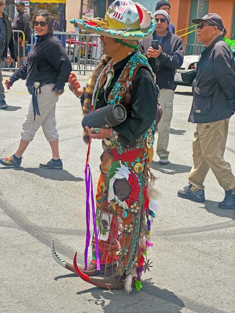 A person in colorful, ornate attire with ribbons, feathers, and animal motifs stands in a street, holding a speaker, surrounded by casually dressed people.
