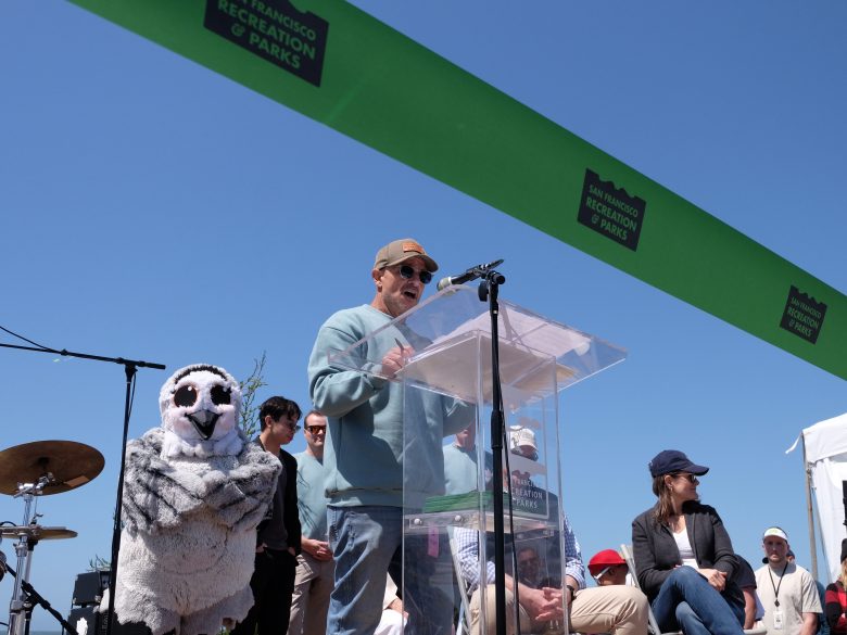 A person speaks at a podium during an outdoor event with a "San Francisco Recreation & Parks" sign; people and a person in an owl costume are nearby under a clear sky.