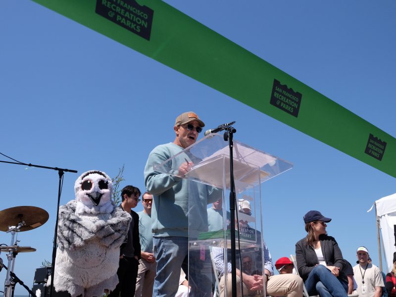 A person speaks at a podium during an outdoor event with a "San Francisco Recreation & Parks" sign; people and a person in an owl costume are nearby under a clear sky.
