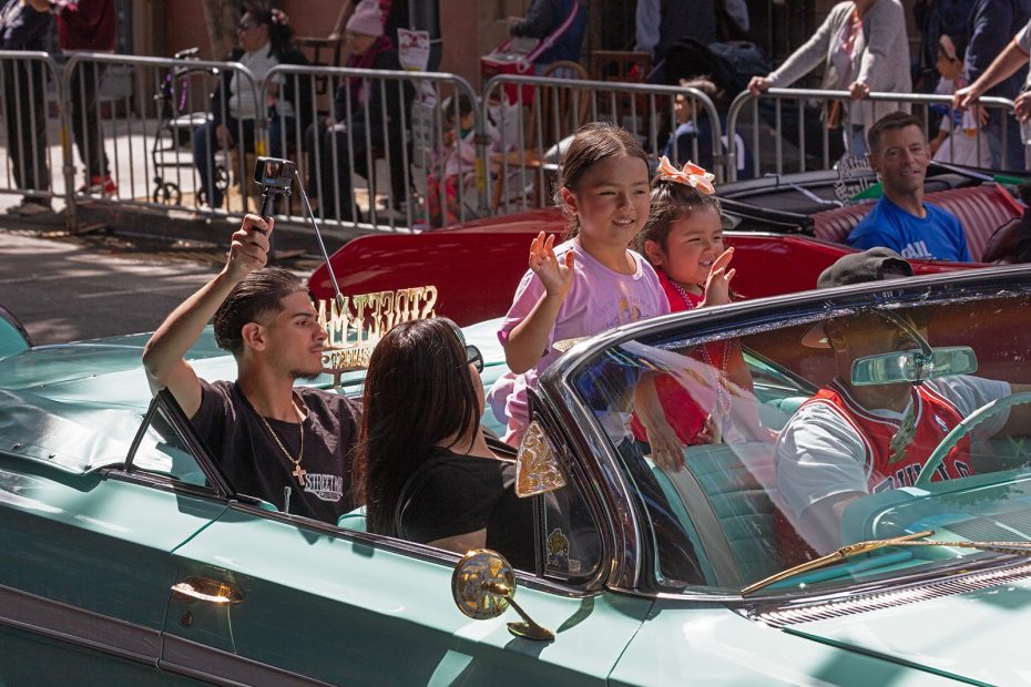 A family rides in a green convertible during a parade; two children stand and wave while an adult passenger films with a phone on a selfie stick.