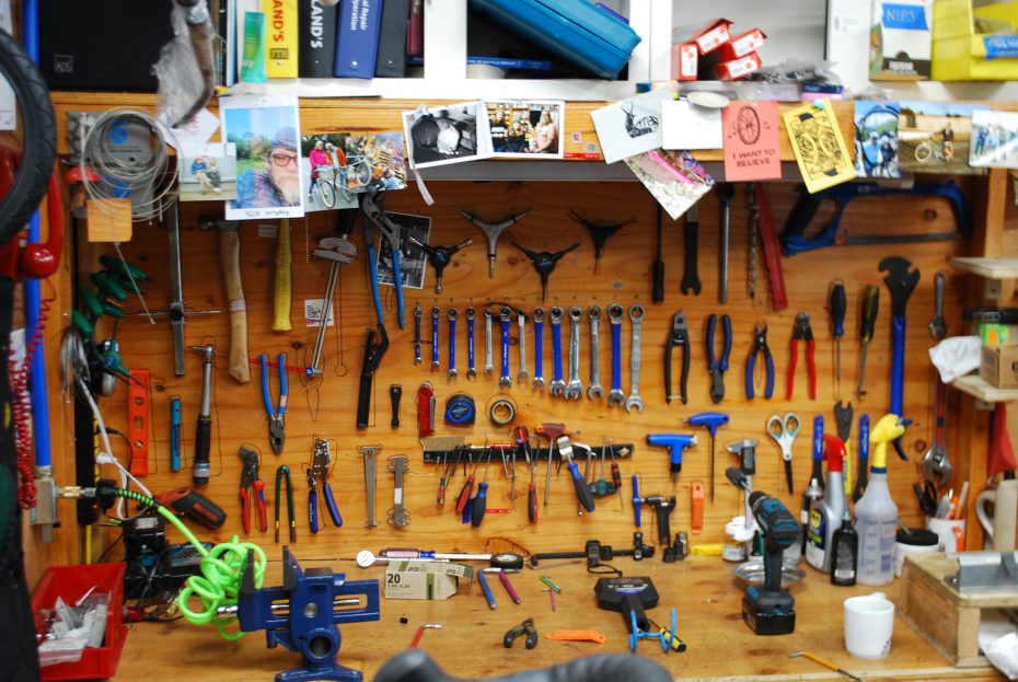 A workbench with various hand tools neatly hanging on a wooden wall, including wrenches, pliers, and screwdrivers, with bottles and supplies on the table and photos pinned above.