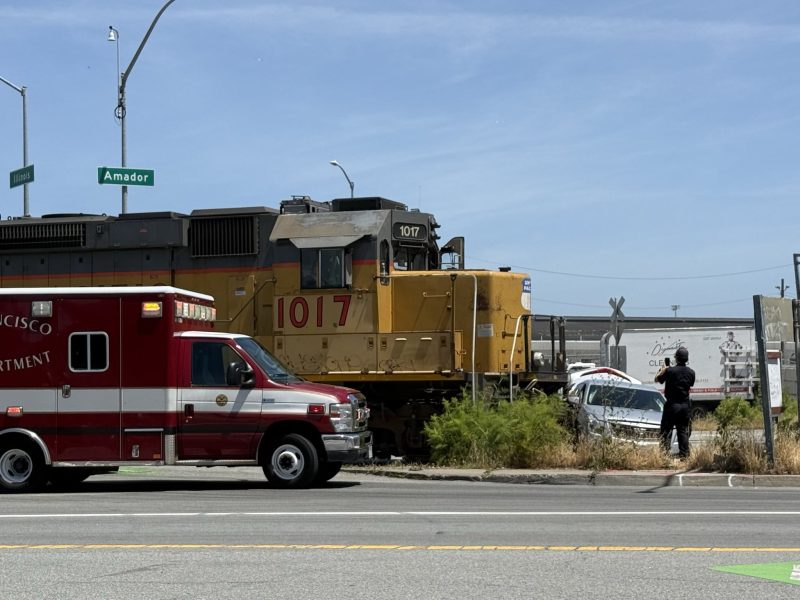 A fire department ambulance, a train, and a damaged car at a roadside scene near an intersection marked "Amador." A person is photographing the area.