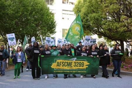 A group of people march outdoors holding an AFSCME 3299 banner and signs advocating for workers' rights; most are wearing green or black shirts.