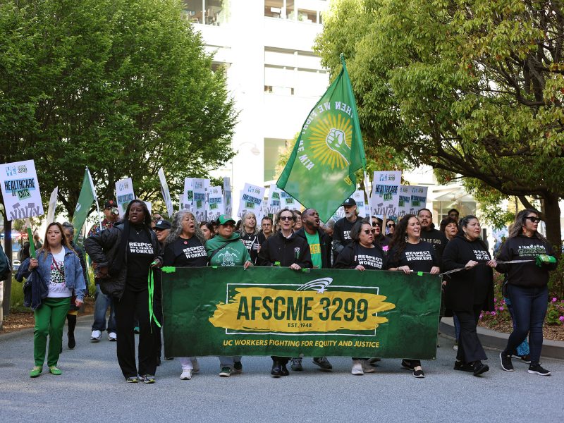 A group of people march outdoors holding an AFSCME 3299 banner and signs advocating for workers' rights; most are wearing green or black shirts.