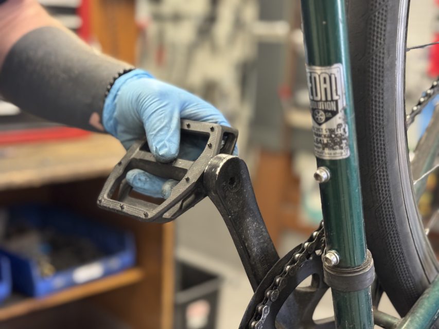 A person wearing a blue glove installs or removes a black bicycle pedal from the crank arm of a green bike.