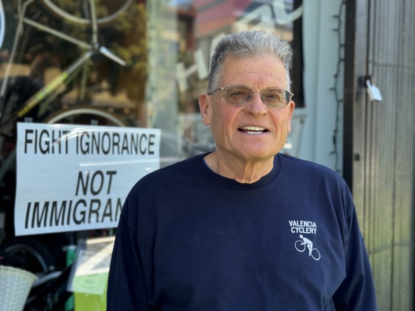 An older man wearing glasses and a Valencia Cyclery shirt stands in front of a window with a sign that reads, “Fight ignorance, not immigrants.”.