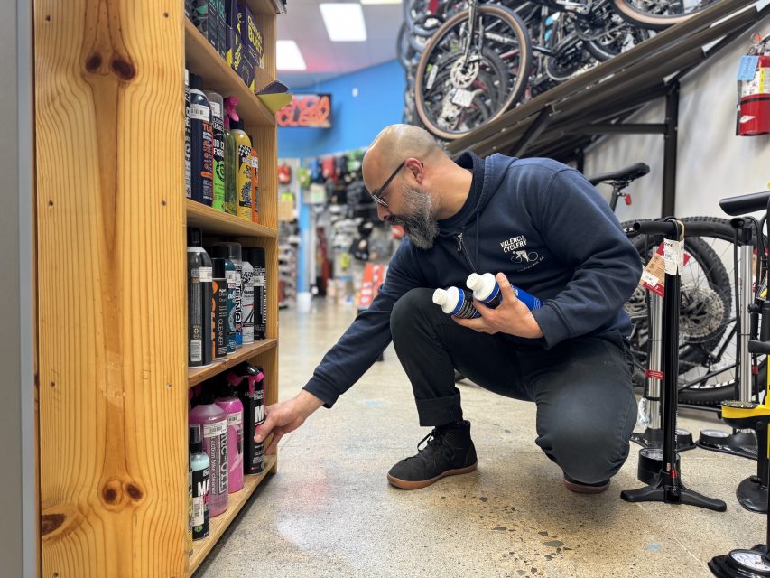 A man kneels in a bike shop, holding bottles while reaching for a product on a lower shelf, surrounded by bike maintenance supplies and bicycles.