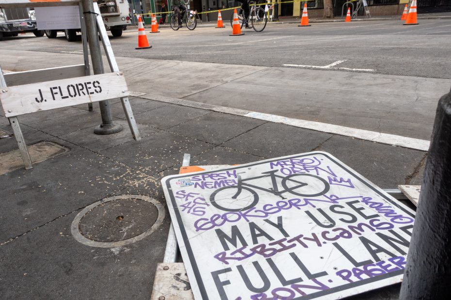 A damaged "May Use Full Lane" bicycle sign with graffiti lies on a city sidewalk near construction cones and a barrier labeled "J. Flores.