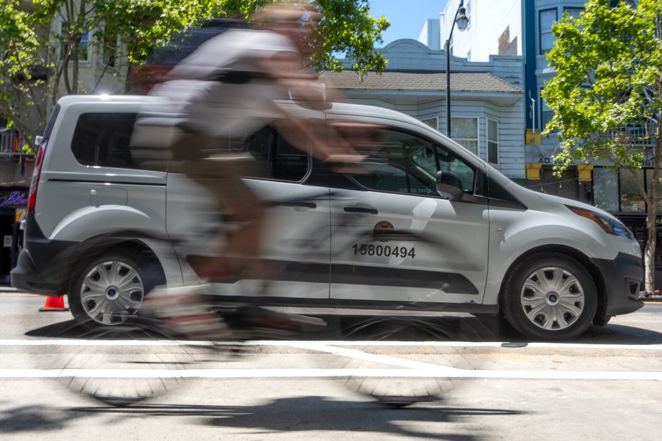 A blurred cyclist rides past a parked white van on a sunny city street with trees and buildings in the background.