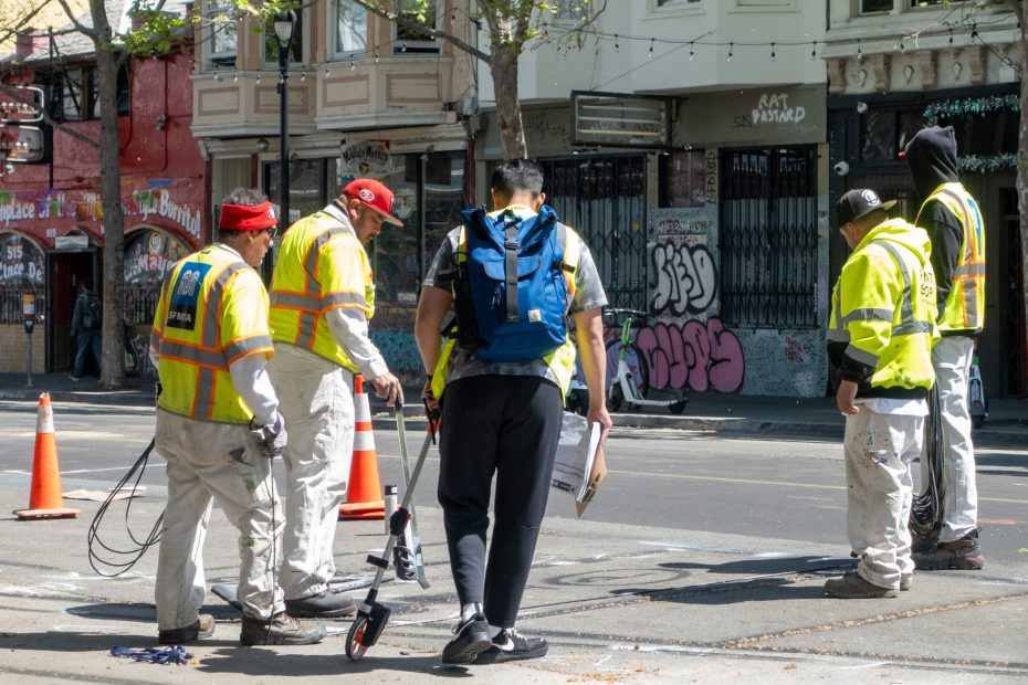 Five workers in reflective vests stand and work on a city street near orange cones; one person is holding a scooter, and graffiti covers the buildings in the background.