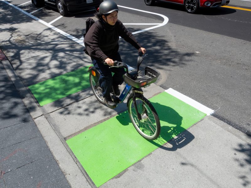A person wearing a helmet rides a bicycle over green-painted markings on a city street bike lane.