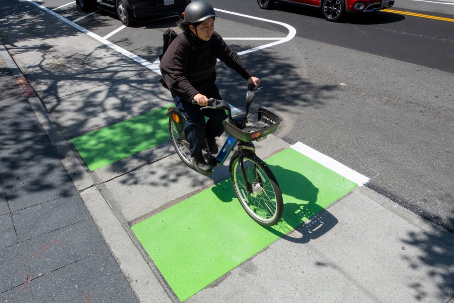 A person wearing a helmet rides a bicycle over green-painted markings on a city street bike lane.