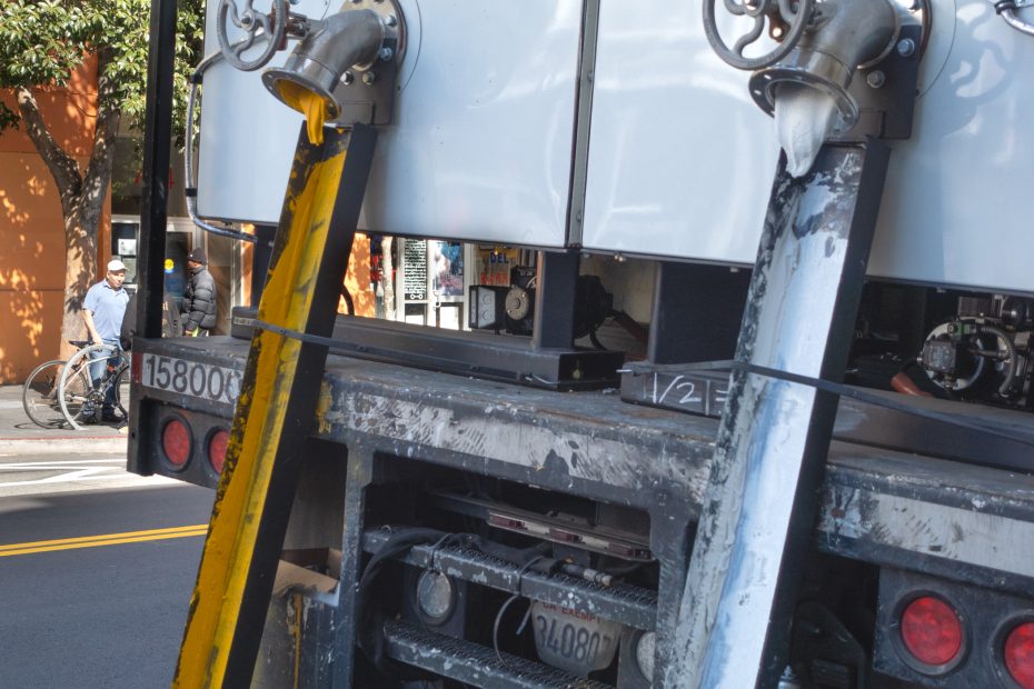 Close-up of the back of a large utility truck with metal equipment and pipes; two people and a bicycle are visible on the sidewalk in the background.