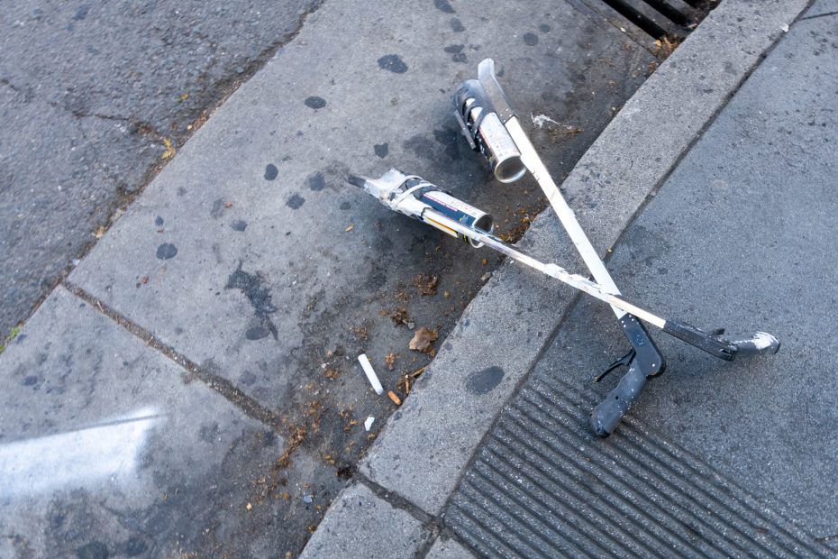 Two crutches lie abandoned on a city sidewalk near the curb, with scattered debris and a storm drain visible nearby.