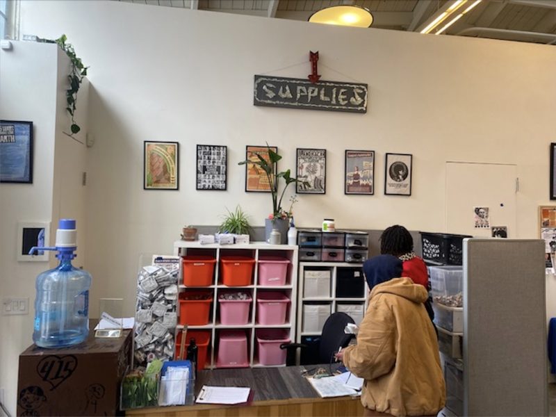Two people stand at a counter in front of cubby shelves with pink and white bins, under a “SUPPLIES” sign and framed posters on a white wall.