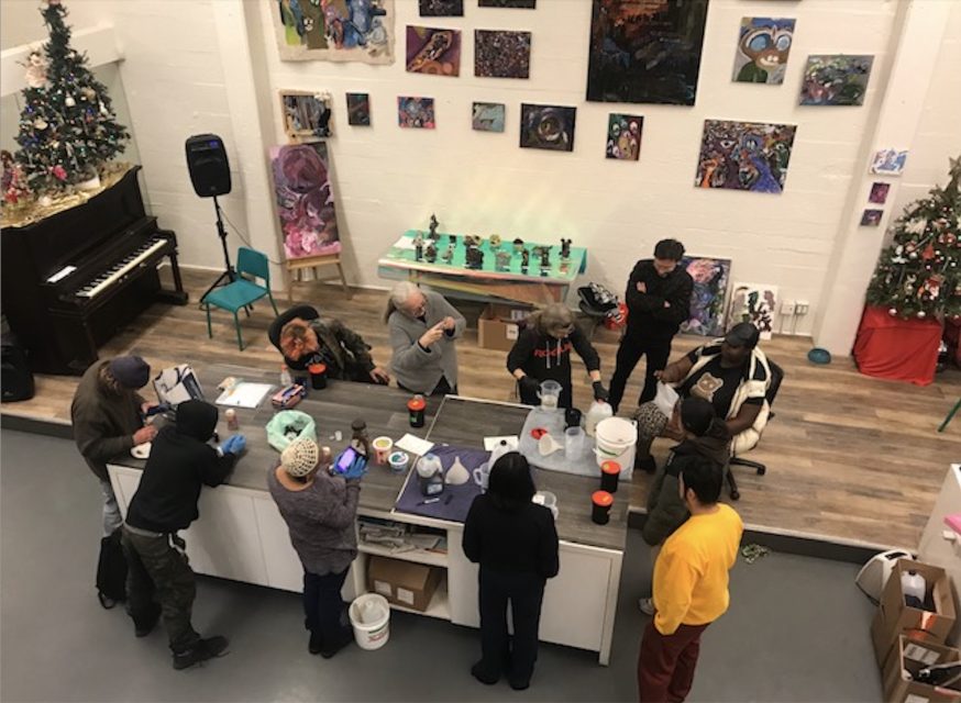 A group of people gather around a kitchen island in an art studio, engaging in activities; artwork and decorated Christmas trees are visible in the background.