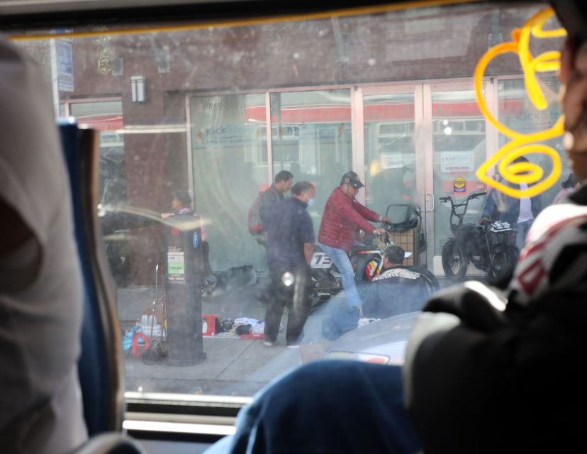 Several people gather around a fallen motorcycle on a city sidewalk, as seen through a bus window; one person appears to be attending to someone on the ground.