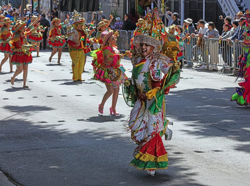People in ornate, colorful costumes dance in a street parade, with onlookers standing behind metal barriers on a sunny day.