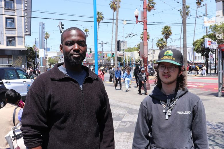 Two men stand side by side on a busy city street with palm trees, pedestrians, and urban buildings in the background.