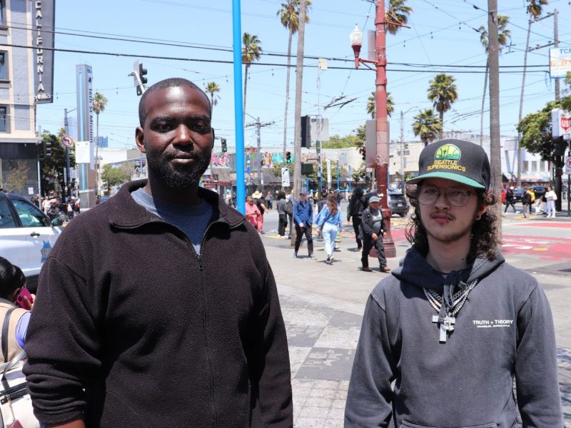Two men stand side by side on a busy city street with palm trees, pedestrians, and urban buildings in the background.