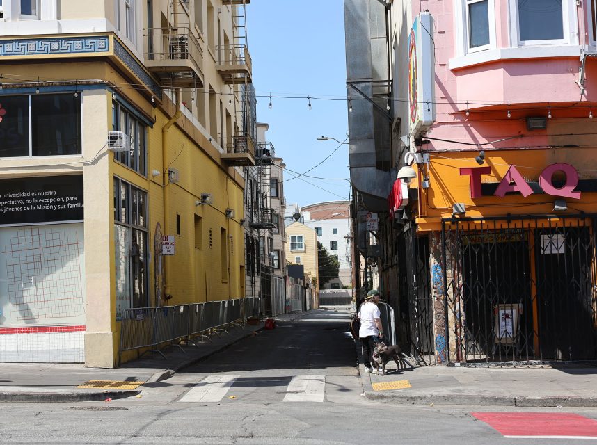 A person walks a dog down a narrow alley between a yellow building and a pink building with a partially visible "TAQ" sign in an urban area.