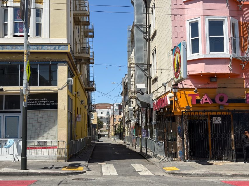 Narrow urban alleyway between two buildings, with yellow and pink facades, a crosswalk in front, and a TAQ restaurant on the right. It is sunny with clear blue skies.
