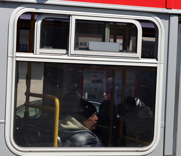 A man wearing a black beanie and plaid jacket sits inside a bus, seen through a window with reflections of the street outside.