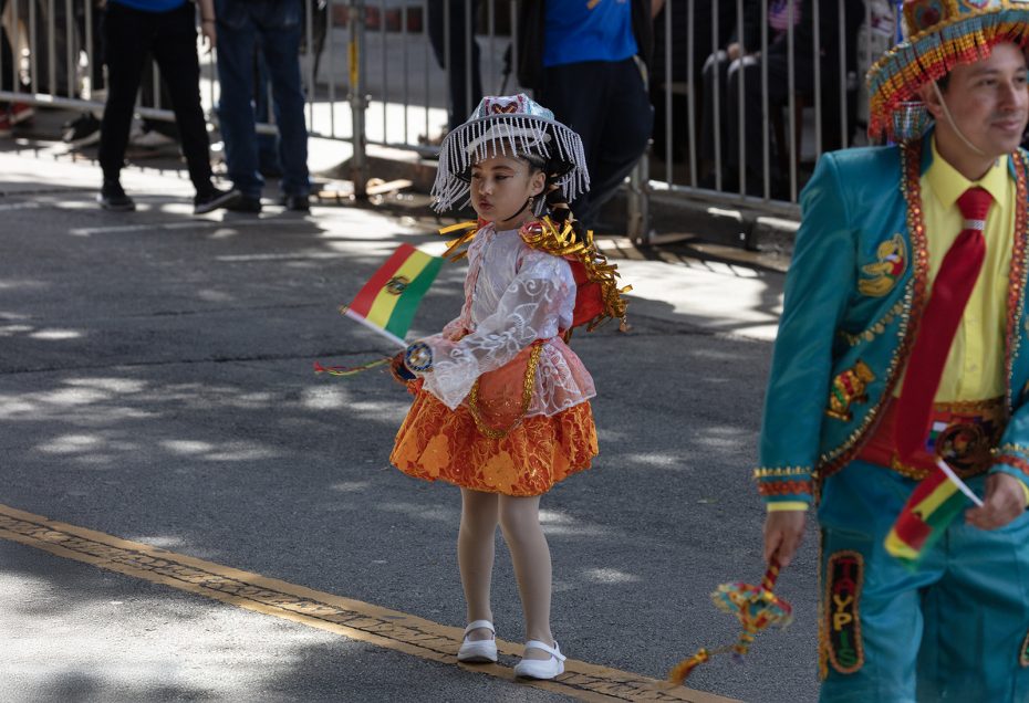 A young girl in a traditional Bolivian costume holds a small flag while standing in the street during a parade, with other participants and onlookers in the background.
