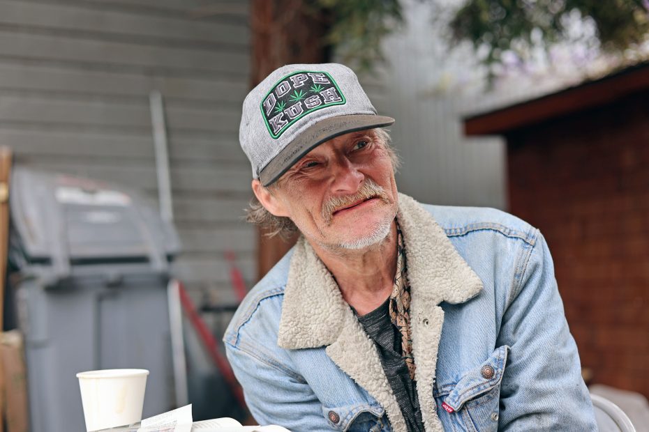An older man wearing a denim jacket and a gray cap with "DOPE KUSH" sits outdoors near a white paper cup and an open book.