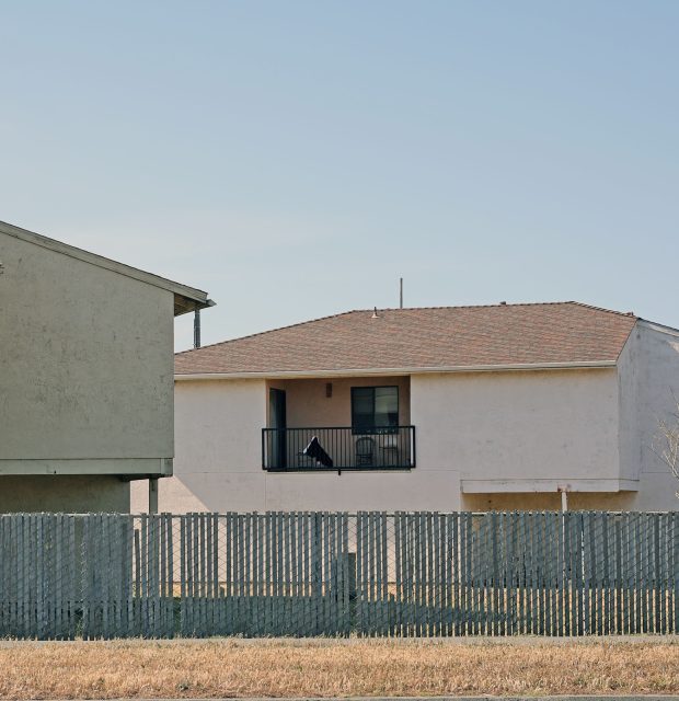 A pale two-story building with a small balcony is partially obscured by a gray wooden fence and another structure under a clear sky.