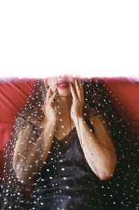 Person in a black lace dress with a sheer polka dot veil, sitting against a red background; upper part of image appears cut off or overexposed.