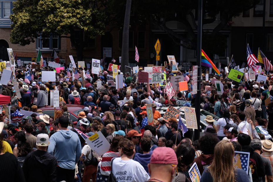 A large crowd gathers outdoors during the day for a protest, holding various signs and flags, including American and rainbow flags.