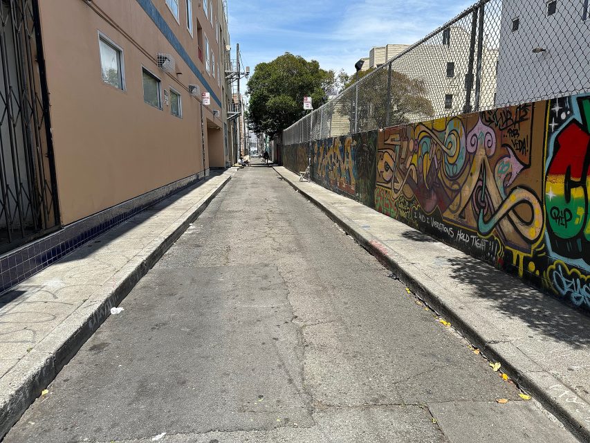 Narrow urban alley with a mural-covered wall on the right, beige buildings on the left, chain-link fence, and clear blue sky overhead.