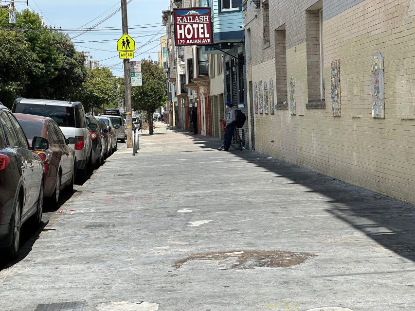 A person sits against a wall near the entrance of Kailash Hotel on Julian Ave; parked cars line the street, and it is a sunny day.