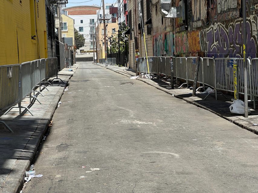 A narrow urban alley lined with metal barricades and scattered trash, bordered by graffiti-covered walls and buildings.