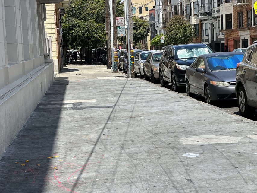 A city sidewalk with parked cars along the street, buildings on both sides, and some scattered litter on the ground in daylight.