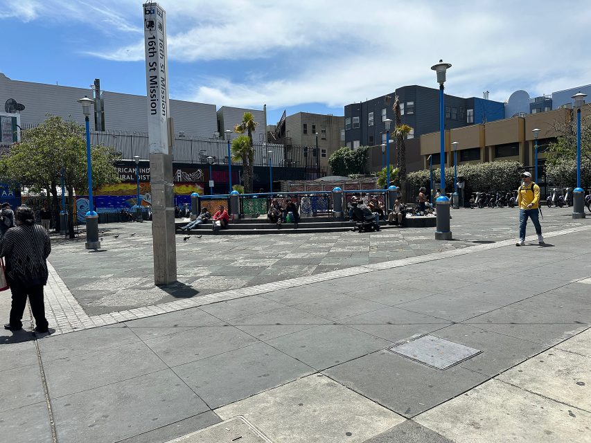 A city plaza with people sitting on benches and steps, a "16th St Mission" signpost, trees, and surrounding buildings under a partly cloudy sky.
