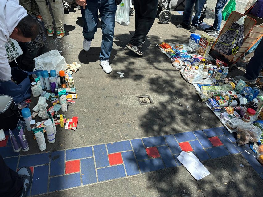 People's legs visible around a sidewalk market displaying toiletries, canned food, and groceries on the ground; partial shadows and a stroller are present.