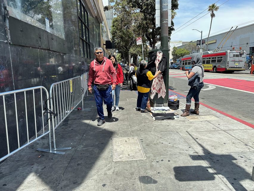 People walk on a city sidewalk beside metal barriers; a few individuals stand near a pole with belongings and blankets, while a bus passes in the street under a sunny sky.
