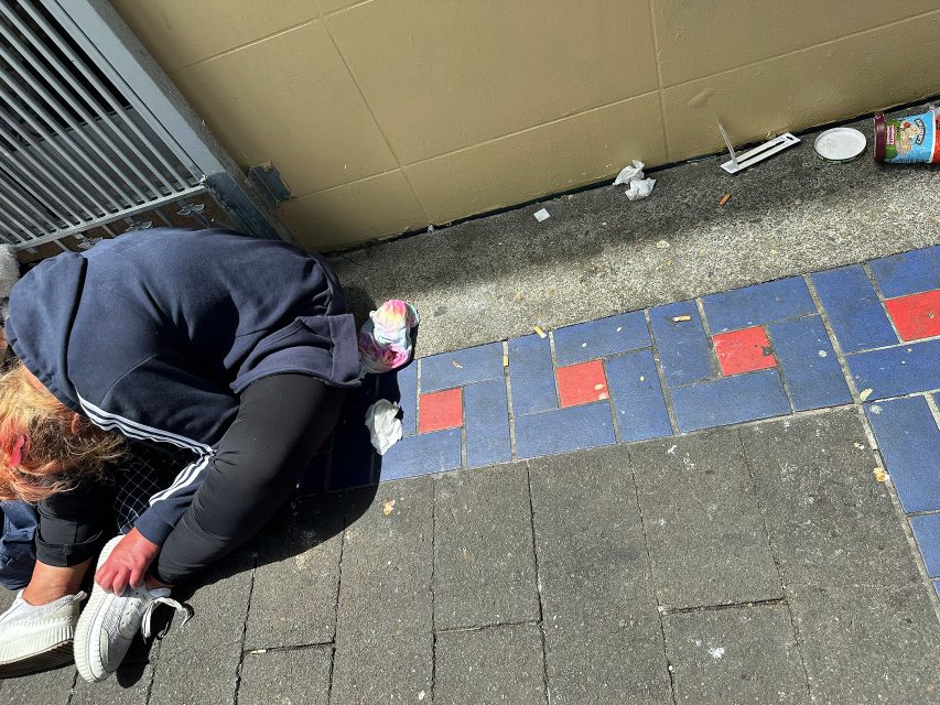A person in dark clothing sits on a sidewalk near a wall, with litter including a cup and scattered trash nearby. Blue and red patterned tiles are visible on the ground.