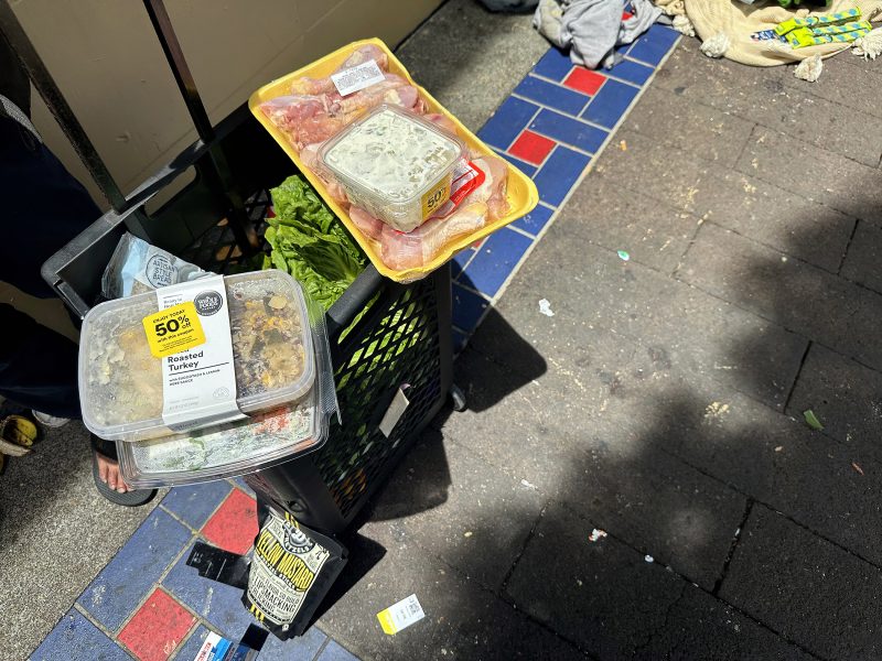 A shopping cart on a sidewalk holds groceries, including packaged chicken, a salad box, and leafy greens, with discount stickers visible on the food items.