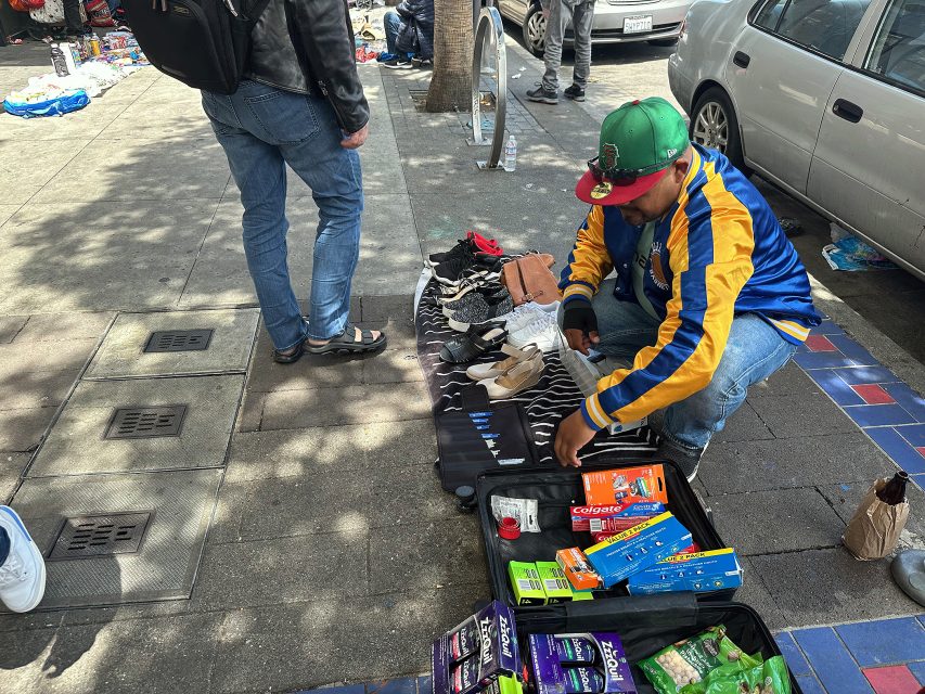 A man in a colorful jacket and green cap arranges toiletries and hygiene products for sale on a sidewalk next to a parked car.