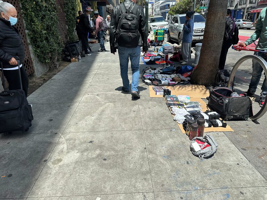 People walk by various items laid out for sale on the sidewalk in an urban setting, with some pedestrians and street vendors visible.
