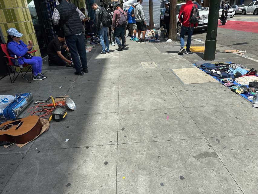 People gather on a city sidewalk with various items, including clothes and a guitar, laid out on the ground for sale or display.