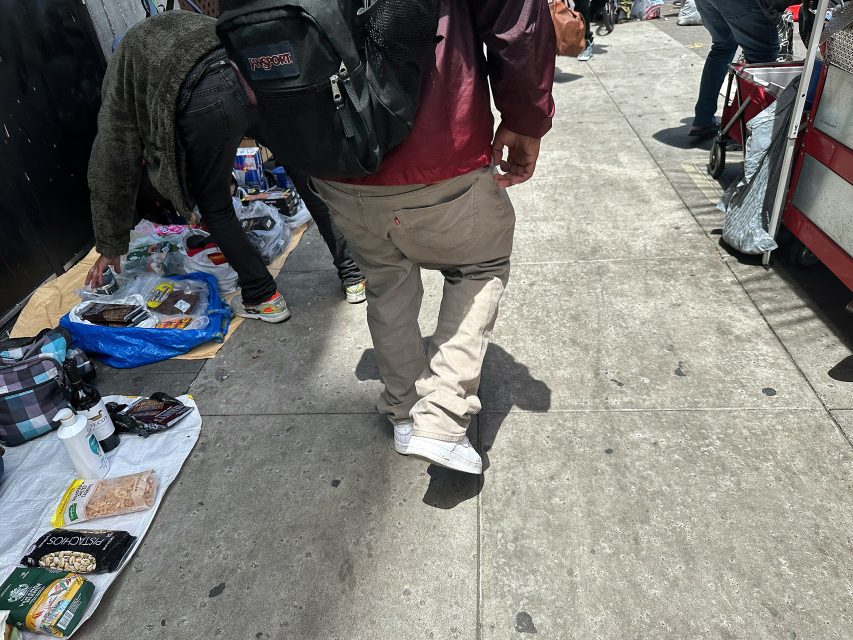 A person with a backpack walks on a city sidewalk past street vendors selling various items laid out on blankets and tarps.